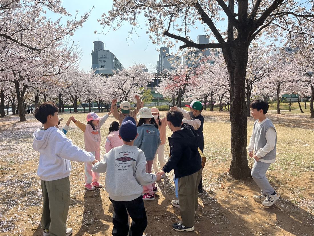 벚꽃이 핀 나무 아래에서 아이들이 손을 잡고 둥글게 서 있는 모습